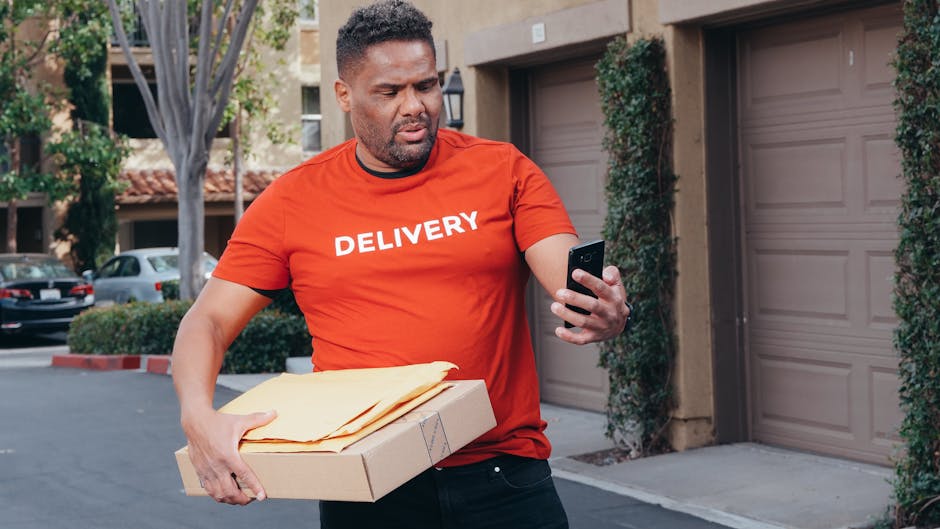 A delivery man looking at his smartphone while holding packages, outdoors.